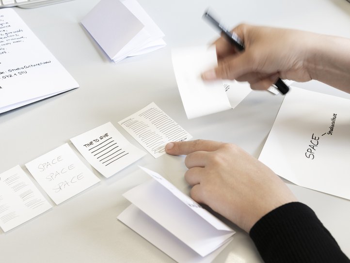 Hände arbeiten an handschriftlichen Notizen an einem Schreibtisch in der 2D-Werkstatt der HfK Bremen. Zu sehen sind kleine Papierhefte mit Texten wie „SPACE“ und „TIME TO LOVE“, die den kreativen Prozess und die Organisation betonen.