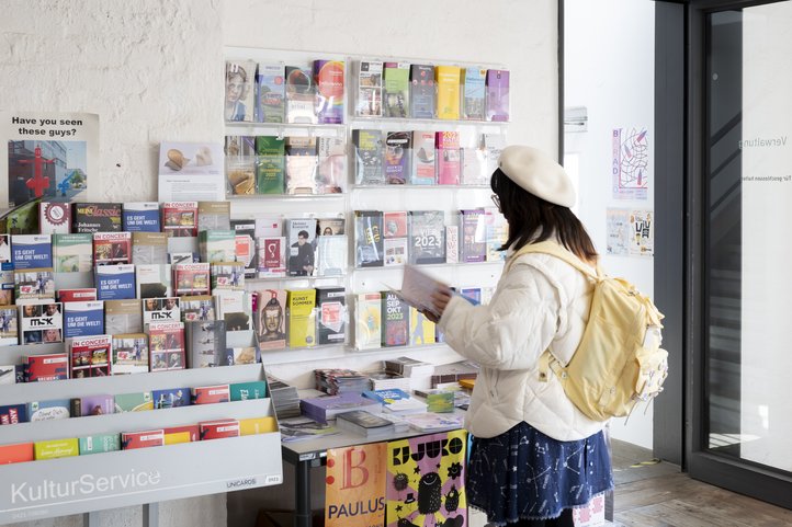 Person in a white jacket and beret stands in front of a display of brochures at a cultural service center, holding a brochure. Display includes arts and event information. Brightly colored posters and pamphlets are visible.