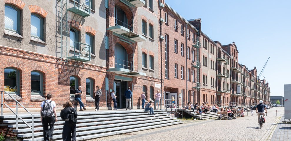 Exterior view of Speicher XI at HfK Bremen, showcasing historic brick architecture with students and visitors interacting on steps and pathways, under a clear blue sky.