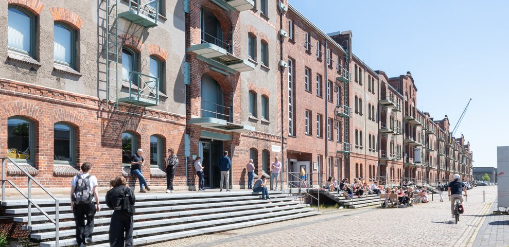 Exterior view of Speicher XI at HfK Bremen, showcasing historic brick architecture with students and visitors interacting on steps and pathways, under a clear blue sky.