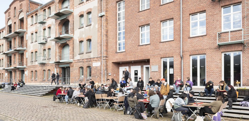 Students sitting at tables in front of the cafeteria in the Speicher building of HfK Bremen, brick building in the background.