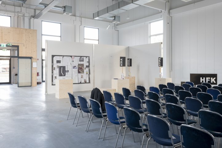 Interior view of Speicher XI A at HfK Bremen featuring rows of blue chairs, wooden partitions with displayed materials, and large windows casting natural light.