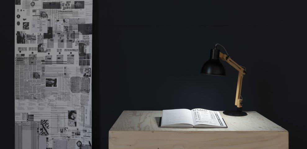Desk lamp illuminating an open book, with a wall full of black-and-white newspaper and document clippings in the background.