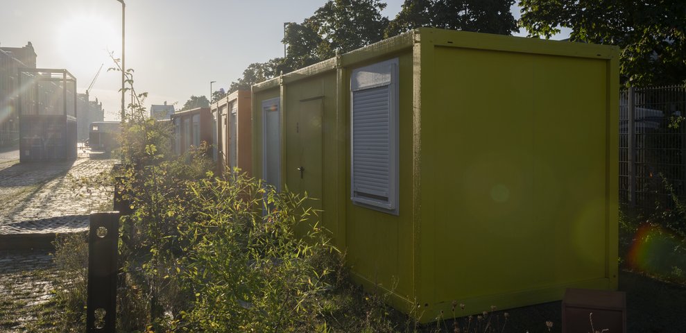 Sunrise over the container village at HfK Bremen with yellow and orange containers, plants in the foreground.