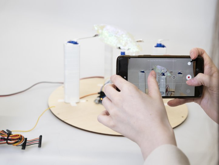 Eine Person fotografiert ein elektronisches Experiment mit einem Smartphone, das verschiedene Drähte und Komponenten zeigt.