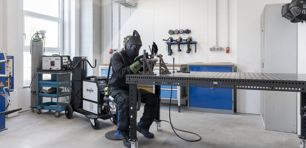 Eine Person arbeitet mit einem Schweißgerät an einem Arbeitsplatz in der Metallwerkstatt der HfK Bremen. Sie trägt Schutzkleidung und ist von Werkzeugen und Ausrüstung umgeben.