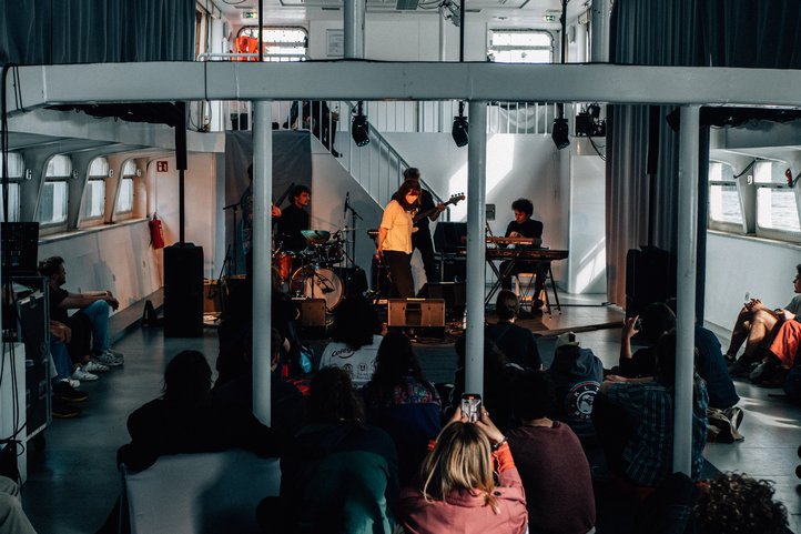 Concert on MS Dauerwelle with band on stage, audience sitting on the floor.