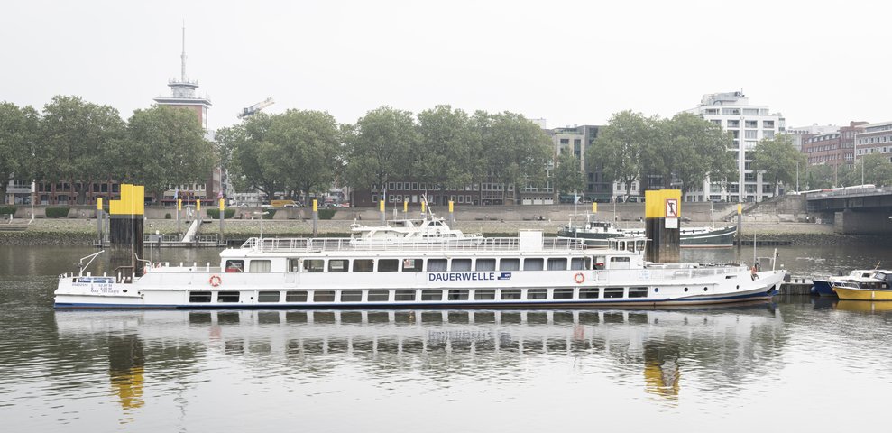 MS Dauerwelle, an event and exhibition ship of HfK Bremen, docked at a riverside with urban buildings and trees in the background.