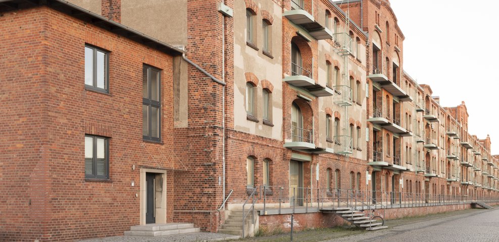 Historic brick facade of Speicher XI at HfK Bremen, showcasing its industrial architectural design and multiple balconies along a cobblestone pathway.