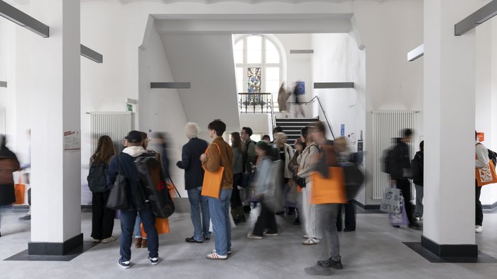 Menschen in Bewegung im Gebäude Dechanatstraße, versammeln sich und gehen mit orangefarbenen Taschen in einem hellen Flur herum.