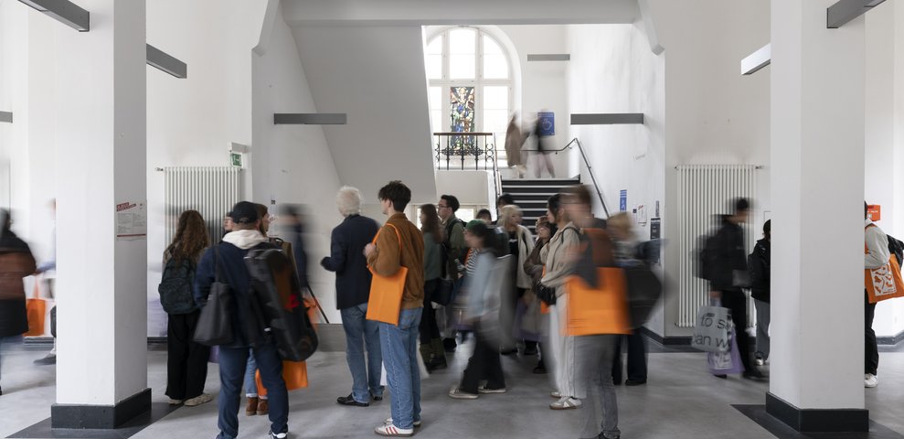 People in motion inside Dechanatstraße building, gathering and walking around with orange bags in a bright hallway.