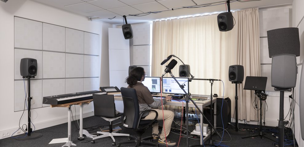 Audiolab at HfK Bremen featuring a person working at a computer with audio equipment, surrounded by speakers, a keyboard, and microphones.