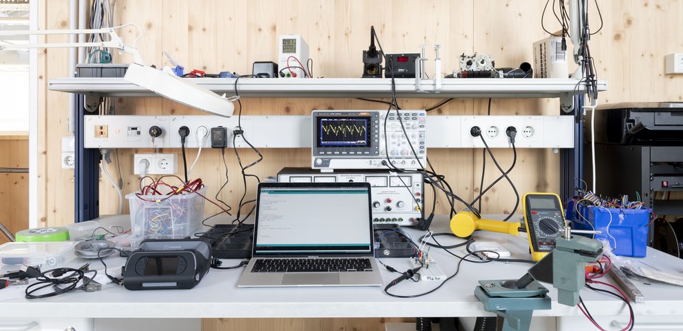Electronics workstation at HfK Bremen featuring an oscilloscope, laptop displaying code, multimeter, wiring tools, and various electronic components.