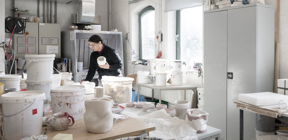 Ceramics Workshop at HfK Bremen with an individual handling ceramics, surrounded by pots, tools, and shelves, well-lit by natural light.