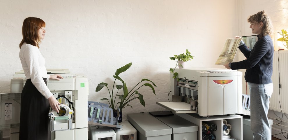 Zwei Personen bedienen Druckmaschinen im Druckpool der HfK Bremen, Pflanzen im Hintergrund.
