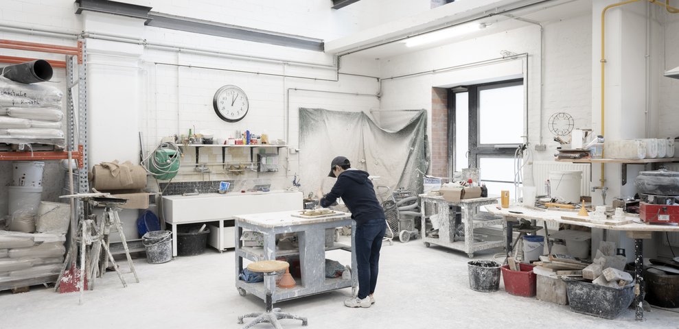 An individual works in the Foundry and Chasing Workshop at the University of the Arts Bremen, surrounded by tools, materials, and equipment in a spacious, well-lit studio.